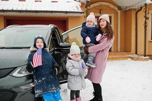 la joven madre americana con hijos sostiene banderas de estados unidos y carga un coche eléctrico en el patio de su casa en invierno. foto