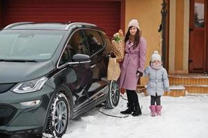 Young woman with baby girl hold eco bags and charging electric car in the yard of her house . photo