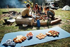 Family bonfire in mountain. Frying sausages. Mother with four kids camping. Autumn hike and camp weather. Warming and cooking near flame together. photo