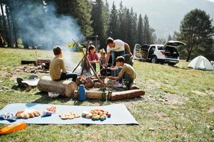 Family bonfire in mountain. Frying sausages. Four kids camping. Autumn hike and camp weather. Warming and cooking near flame together. photo