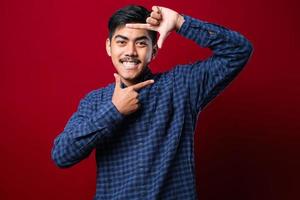 Young handsome asian boy wearing casual shirt standing over isolated red background smiling making frame with hands and fingers with happy face. photo