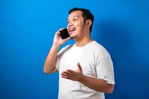 Fat asian man in white t-shirt look happy while talking on smartphone. Half body studio shots against blue background photo