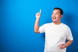 retrato de un hombre asiático gordo y divertido con una camiseta blanca sonriendo y bailando alegremente, expresando alegría celebrando la victoria de las buenas noticias ganando el gesto de éxito contra el fondo azul foto