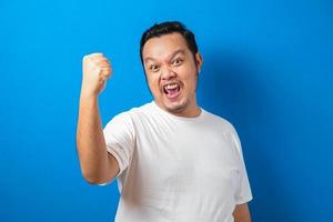 Portrait of a funny fat Asian man in white t-shirt smiling and dancing happily, joyful expressing celebrating good news victory winning success gesture against blue background photo