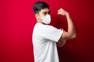 Young asian man in white t-shirt wearing medical mask standing over isolated red background showing arms muscles smiling proud. photo