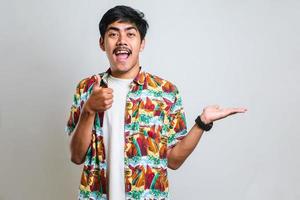 Asian man in white tshirt smiling and pointing to presenting something on his side, against red background with copy space photo