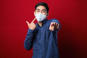 Portrait of young Asian student wearing flannel shirt and mask pointing forward, looking at camera choosing someone to wear a mask photo