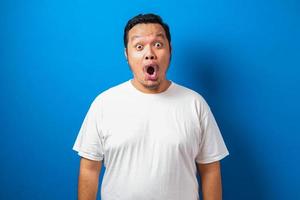 un hombre asiático gordo con una camiseta blanca muestra una expresión graciosa y sorprendida contra el fondo azul foto