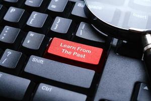 Selective focus of magnifying glass on a computer keyboard with red button written with Learn From Past photo