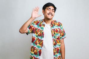Man wearing white tshirt standing over white background Waiving saying hello happy and smiling, friendly welcome gesture photo