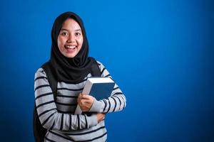 estudiante universitaria musulmana asiática usando hijab sonriendo amigable sosteniendo un libro grande foto