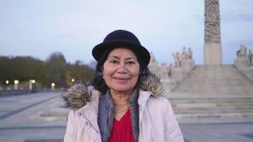 vue de face d'une femme asiatique debout, souriante et regardant la caméra dans le parc vigeland, norvège. belle journée avec de belles sculptures et ciel bleu. video