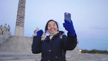 gelukkige aziatische vrouw die staat en een videogesprek aanneemt in vigeland park, noorwegen. mooie dag met prachtige sculpturen en blauwe lucht, oslo video