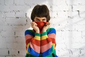 A young woman in a bright multicolored rainbow sweater hides her face and covers her ears with her hands. photo