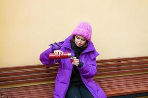 A young woman in warm outerwear pours hot tea from a thermos while sitting photo