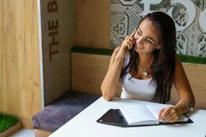 Positive woman talking on the phone in a cafe with an open notebook. Caucasian photo
