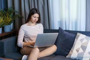 Young woman sitting on couch, working at laptop, browsing internet or reading photo