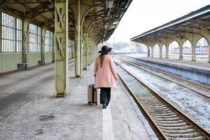 Woman in pink coat walks with suitcase on an empty platform with her back
