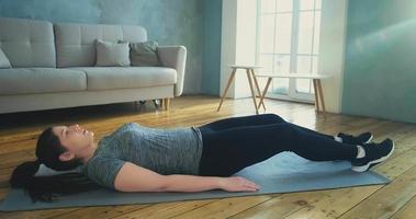 jonge langharige vrouw in stijlvolle sportkleding doet crunches en beenliften op grijze mat in woonkamer met parket slow motion video