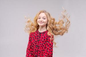 A beautiful cheerful girl in red dress with flying long wavy hair on light gray photo