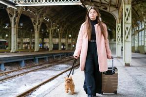 Travel concept. At station, young tourist with dog goes and drags suitcase photo