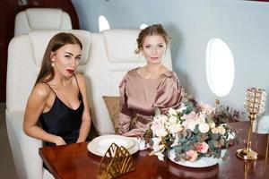 dos hermosas mujeres con estilo están sentadas a la mesa en un jet privado. celebrando foto