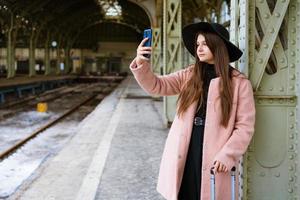 mujer joven feliz en la plataforma de la estación de tren con abrigo rosa y sombrero negro foto