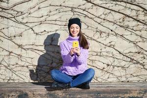 Happy young woman smiling sitting on a bench outdoors using a smartphone. photo