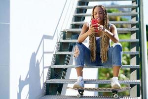 mujer negra con trenzas de colores, consultando su teléfono inteligente con los pies apoyados en una patineta. foto