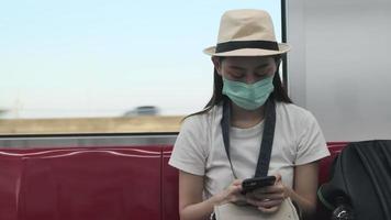 Face mask Asian female tourist with hat sits in sky train, message chatting mobile phone, travel transporting with a public railway, city passenger lifestyle, new normal journey vacation in Thailand. video