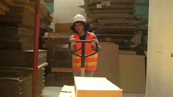 Female warehouse worker in safety uniform and hardhat using hydraulic trolley haul piles of cardboard for shipping and logistic transport at manufacture factory, supply stocks in packaging industrial. video