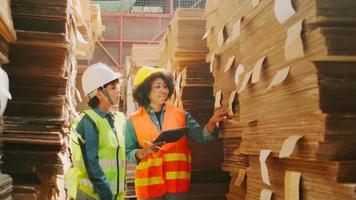 Two female workers and colleagues in safety uniforms and helmet stock check, control production in storage of warehouse factory with a lot of paper stack, friend works in recycle manufacture business. video