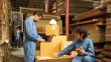 Two safety uniform workers and colleagues use laptop and mobile phone to check shipment orders stock at parcels warehouse, paper manufacture factory for packing industry, logistic transport service.