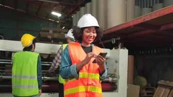 African American female worker in safety uniform and hard hat chatting mobile phone, looking to camera and smile, mechanical supervisor in industrial machine of paper production manufacture factory.