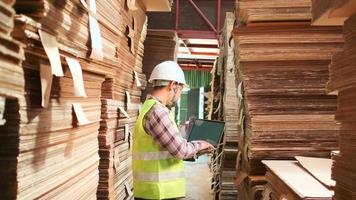 Asian male worker in safety uniform and hard hat, supervisor inspector with laptop for packaging stock order at factory warehouse, piles of stacking paper manufacture, recycling production industry. video