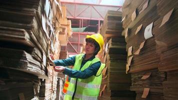 Female worker in safety uniform and hard hat, supervisor quality inspects packaging stock order supply at factory storage warehouse, piles of stacking paper manufacture, recycling production industry.