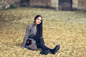 Beautiful girl wearing winter coat sitting on the floor of an urban park full of autumn leaves. photo