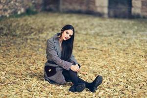 hermosa chica con abrigo de invierno sentada en el suelo de un parque urbano lleno de hojas de otoño. foto