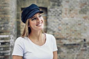 Young blonde woman wearing cap smiling near a brick wall. photo