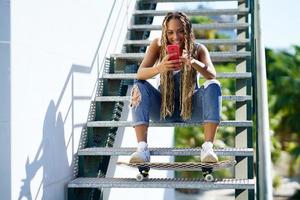 Black woman with coloured braids, consulting her smartphone with her feet resting on a skateboard. photo