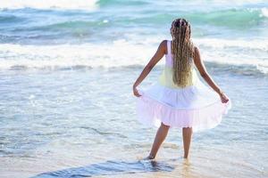 chica negra caminando por la orilla de la playa con un hermoso vestido largo. foto