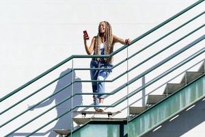 mujer negra con trenzas de colores, mirando su teléfono inteligente con los pies apoyados en una patineta. foto