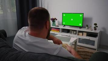 hombre viendo la clave de croma de tv de pantalla verde. vista trasera de un hombre de mediana edad sentado en un sofá relajante sala de estar en la televisión. video