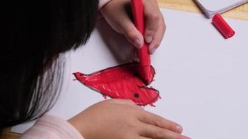 close-up van een klein meisje dat een hart tekent en kleurt met haar gekleurde stiften. schattig jong meisje huiswerk aan tafel thuis. kunsten en ambachten. video