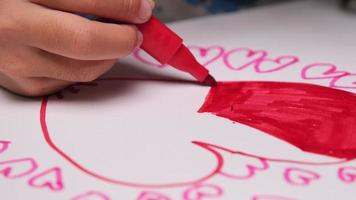 gros plan d'une petite fille dessinant un coeur et coloriant avec ses marqueurs de couleur. jolie jeune fille faisant ses devoirs à table à la maison. l'artisanat. video