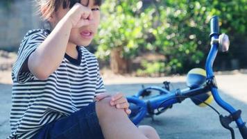 niña triste sentada en el suelo después de caerse de su bicicleta en el parque de verano. niño resultó herido mientras montaba en bicicleta.