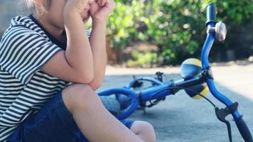 niña triste sentada en el suelo después de caerse de su bicicleta en el parque de verano. niño resultó herido mientras montaba en bicicleta. video