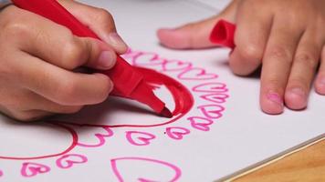 närbild av en liten flicka som ritar ett hjärta och färglägger med sina färgade markörer. söt ung flicka gör läxor vid bordet hemma. konst och hantverk. video