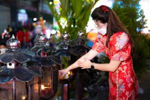 Beautiful woman wearing red cheongsam is filling lamp with oil to keep light on. Beliefs about making merit. Person wearing white face mask. photo