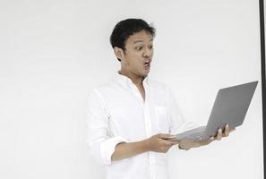 Wow face of Young Asian man shocked what he see in the laptop when working isolated grey background wearing white shirt photo